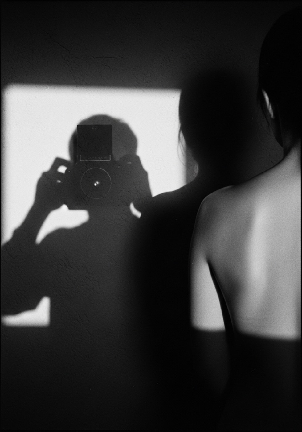 a sombra da fotógrafa com a câmera na parede, ao lado a curva desfocada de umas costas