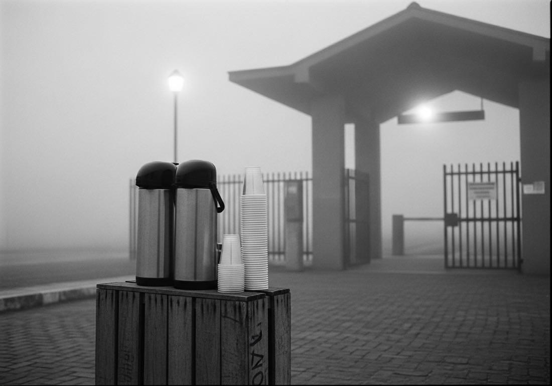 Garrafas térmicas e copinhos sobre caixote de madeira na porta de uma estação de trem, na neblina da madrugada